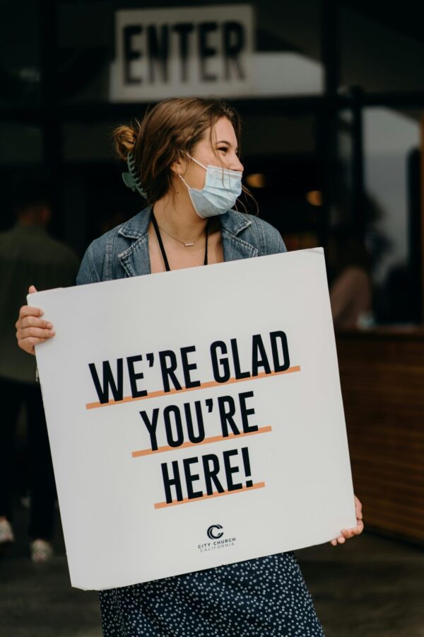 Sign welcoming people to an inclusive church proclaiming Progressive Christianity Woman holding sign that reads, "We're Glad You're Here!" pointing to Progressive Christianity's embrace of everyone.